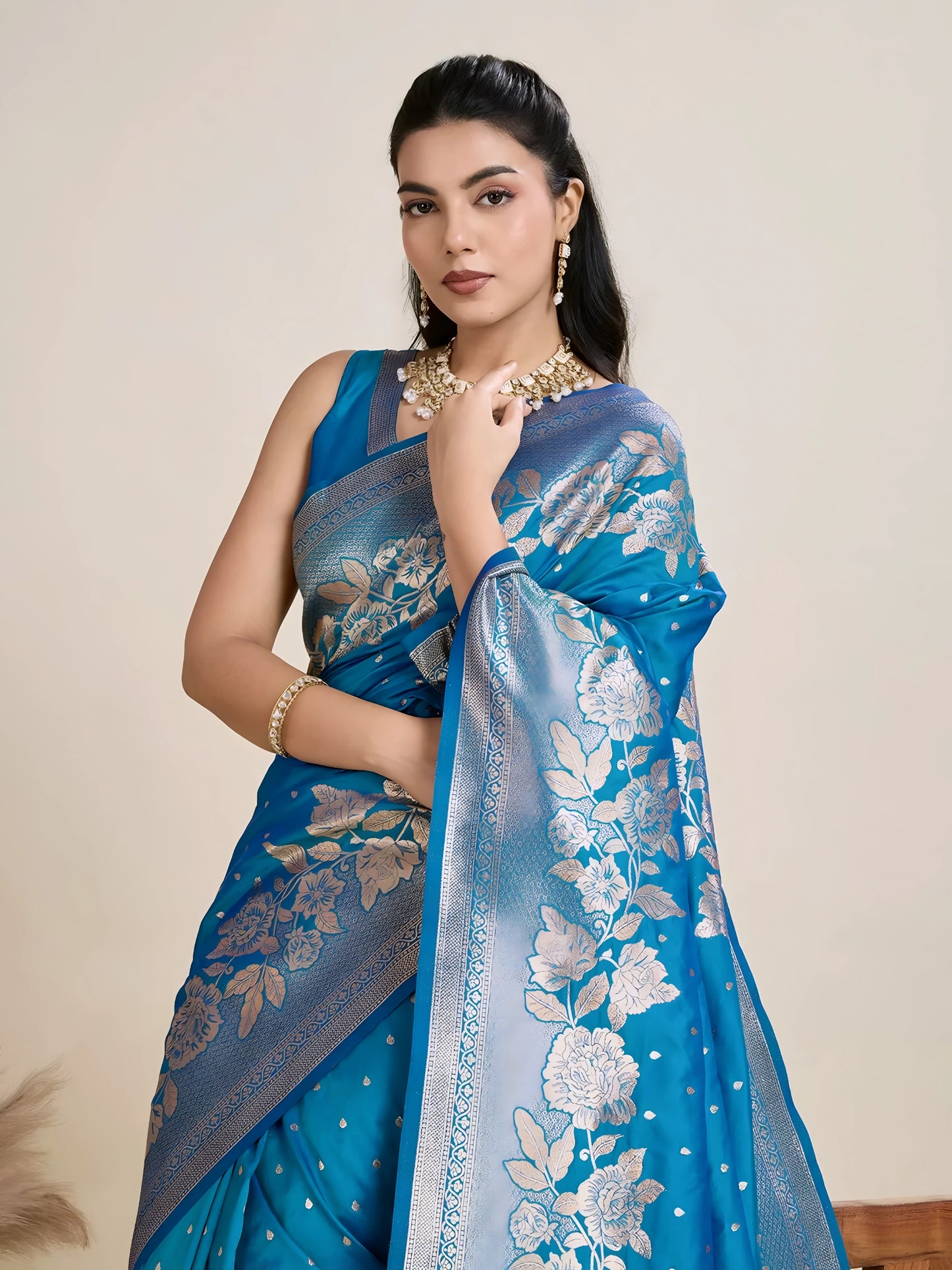 Woman wearing a vibrant blue silk saree with silver floral zari border and pallu, paired with a matching blouse and traditional jewelry, elegantly posing beside a wooden chair in a minimalist studio with neutral decor and ceramic vases.