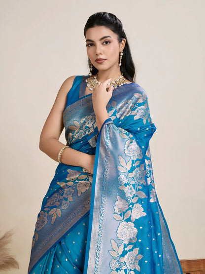 Woman wearing a vibrant blue silk saree with silver floral zari border and pallu, paired with a matching blouse and traditional jewelry, elegantly posing beside a wooden chair in a minimalist studio with neutral decor and ceramic vases.