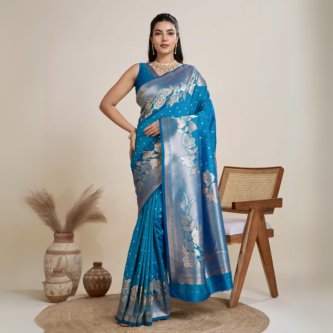 Woman wearing a vibrant blue silk saree with silver floral zari border and pallu, paired with a matching blouse and traditional jewelry, elegantly posing beside a wooden chair in a minimalist studio with neutral decor and ceramic vases.