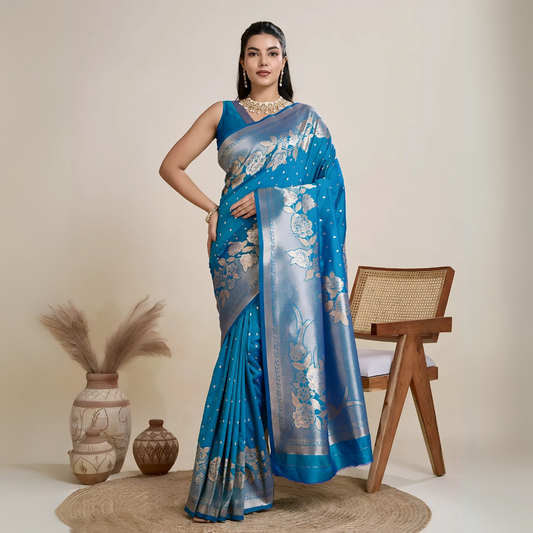 Woman wearing a vibrant blue silk saree with silver floral zari border and pallu, paired with a matching blouse and traditional jewelry, elegantly posing beside a wooden chair in a minimalist studio with neutral decor and ceramic vases.