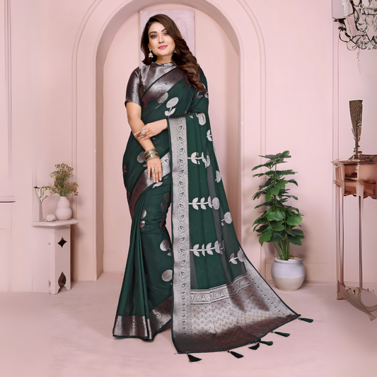 Woman wearing a bottle green silk saree adorned with large silver woven motifs and an intricate silver zari border, paired with a metallic silver blouse. She stands elegantly in front of a pastel arched alcove in a modern studio, accessorized with traditional bangles, with a potted plant and minimalist decor elements in the background.