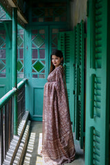 Woman wearing a brown floral printed saree with a matching blouse and statement earrings, standing on a vintage balcony with teal green shutters and doors, bathed in natural light.