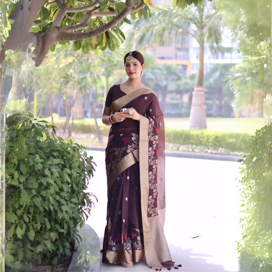 Woman wearing a brown saree with gold border and floral embroidery, posing outdoors under a leafy tree with lush greenery and palm trees in the background.
