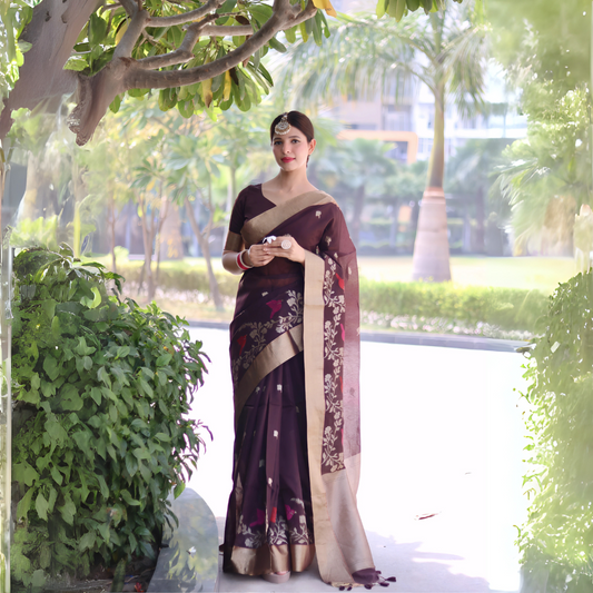 Woman wearing a brown saree with gold border and floral embroidery, posing outdoors under a leafy tree with lush greenery and palm trees in the background.