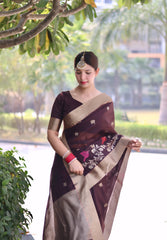 Woman wearing a brown saree with gold border and floral embroidery, posing outdoors under a leafy tree with lush greenery and palm trees in the background.