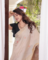 A woman models a Soft Muga Cotton Saree. The saree is light beige/cream, transitioning to a brown/rust-colored pallu featuring an elaborate, colorful Resham and Zari woven design of a tree and birds. She wears a black ruffled blouse and an oxidized silver necklace.