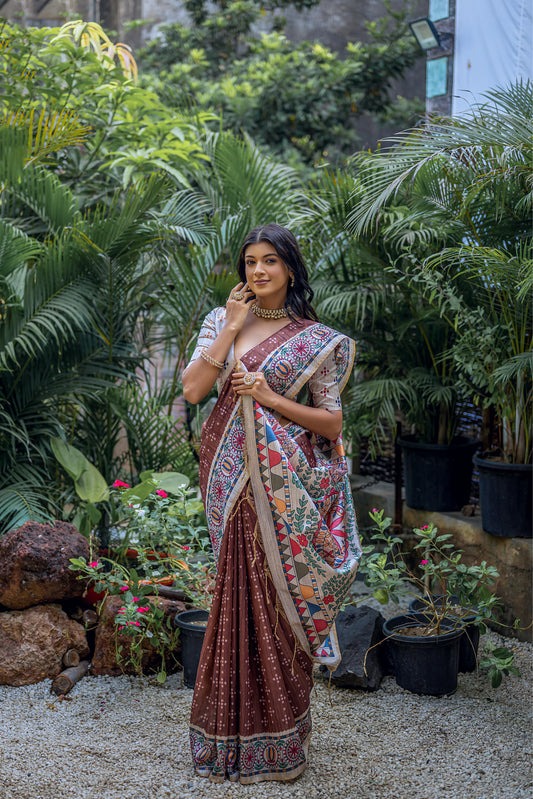 A woman models a deep Brown Tussar Silk Saree with small scattered prints. The pallu features a wide, elaborate panel with intricate Madhubani-style folk art prints in white, red, blue, and green. She wears a cream/gold embellished blouse and a gold Kundan choker necklace.