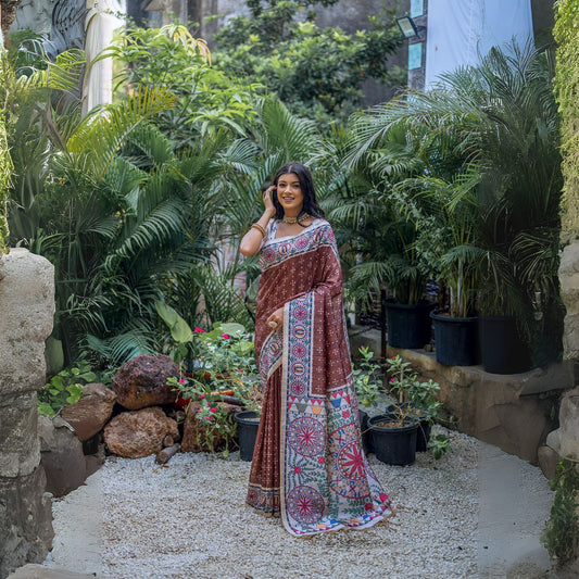 Woman wearing a brown saree with intricate white tribal-inspired prints and a multicolor pallu featuring circular and geometric patterns, accessorized with traditional jewelry, standing gracefully in a lush outdoor garden surrounded by tropical plants.