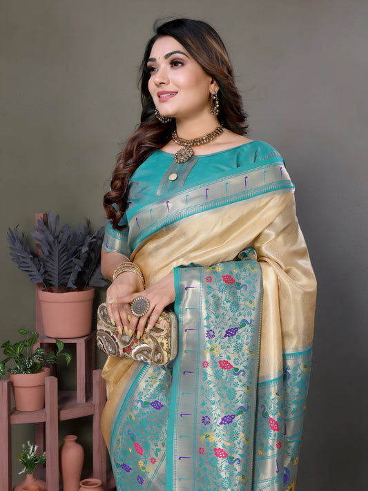 Woman wearing a cream silk saree with a sky blue border and multicolor floral motifs, paired with a matching blue blouse and traditional jewelry. She poses gracefully in a modern studio with minimalist decor, featuring potted plants and terracotta vases on wooden stands against a muted backdrop.