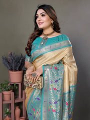 Woman wearing a cream silk saree with a sky blue border and multicolor floral motifs, paired with a matching blue blouse and traditional jewelry. She poses gracefully in a modern studio with minimalist decor, featuring potted plants and terracotta vases on wooden stands against a muted backdrop.