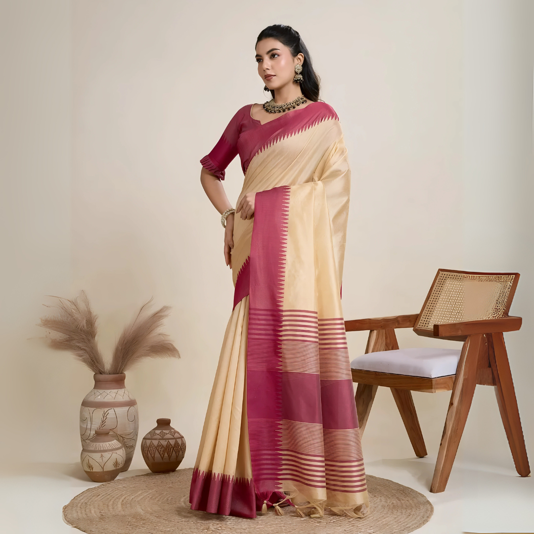 Woman wearing a cream silk saree with a bold maroon geometric border and pallu, paired with a matching maroon blouse and traditional jewelry, elegantly posing beside a wooden chair in a minimalist studio setting with neutral decor, ceramic vases, and pampas grass.
