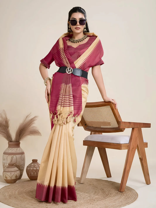 Woman wearing a cream silk saree with a bold maroon geometric border and pallu, paired with a matching maroon blouse and traditional jewelry, elegantly posing beside a wooden chair in a minimalist studio setting with neutral decor, ceramic vases, and pampas grass.