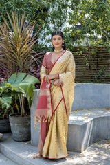 Woman wearing a cream silk saree with maroon border and gold motifs, paired with a matching maroon blouse and traditional jewelry, standing gracefully outdoors on garden steps surrounded by lush green plants and tropical foliage.