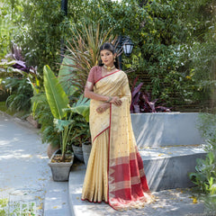 Woman wearing a cream silk saree with maroon border and subtle gold motifs, paired with a matching maroon blouse and traditional jewelry, standing gracefully outdoors amidst lush green plants and garden steps.