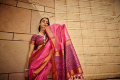 Woman draped in dark pink saree with intricate zari work and ornate pallu, leaning against a stone wall in ethnic attire