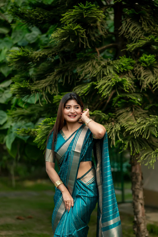 Woman in a dark green saree with zari work standing on a garden pathway surrounded by lush greenery