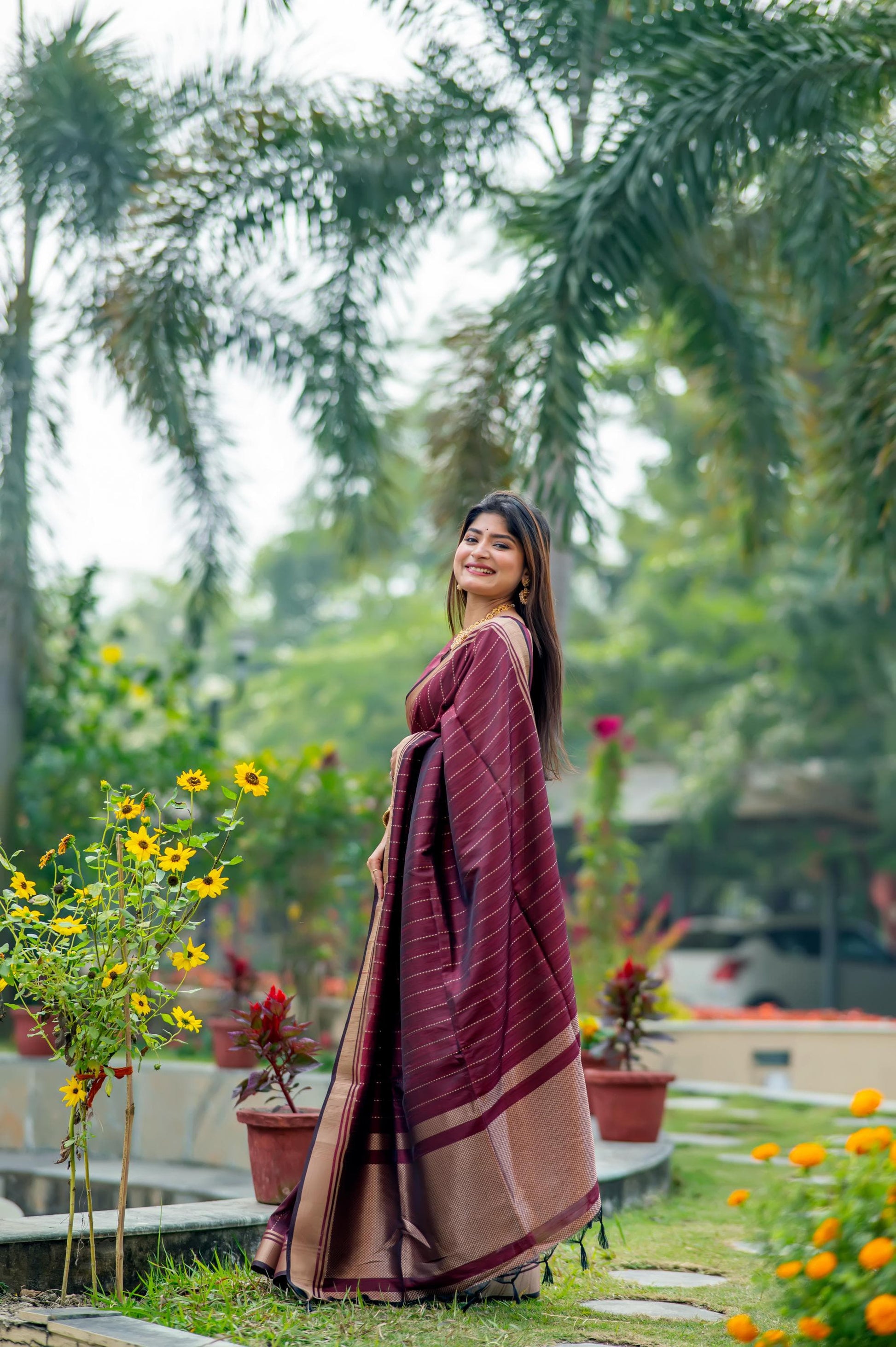 Woman in a deep purple saree with zari work standing in a lush garden with yellow flowers and palm trees in the background