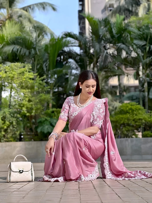 Woman wearing a dusty pink saree with intricate white floral embroidery along the borders and scattered motifs, styled with matching bangles and earrings. She poses outdoors in a garden setting with lush green trees and parked cars in the background, highlighting the saree's elegant and graceful design.