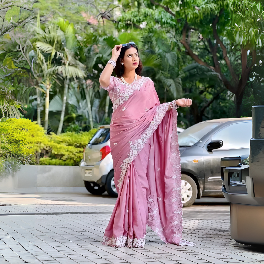 Woman wearing a dusty pink saree with intricate white floral embroidery along the borders and scattered motifs, styled with matching bangles and earrings. She poses outdoors in a garden setting with lush green trees and parked cars in the background, highlighting the saree's elegant and graceful design.