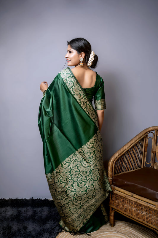 Woman wearing an emerald green silk saree with intricate gold brocade border and pallu, paired with a matching blouse and gold jewelry, standing beside a modern wooden floor lamp with a blue shade in a minimalist studio setting.