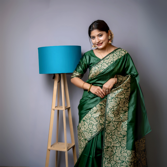 Woman wearing an emerald green silk saree with intricate gold brocade border and pallu, paired with a matching blouse and gold jewelry, standing beside a modern wooden floor lamp with a blue shade in a minimalist studio setting.