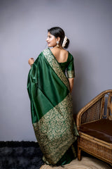 Woman wearing an emerald green silk saree with intricate gold brocade border and pallu, paired with a matching blouse and gold jewelry, standing beside a modern wooden floor lamp with a blue shade in a minimalist studio setting.