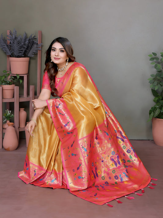 Woman wearing a golden silk saree with a vibrant pink floral embroidered border and multicolor motifs, paired with a matching pink blouse and gold jewelry including a choker necklace, bangles, and rings. She poses gracefully in a modern studio decorated with potted green plants and pastel terracotta vases on wooden stands against a neutral background.