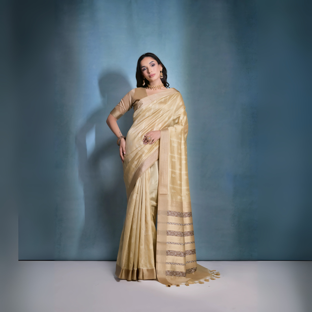 Woman wearing a golden silk saree with intricate border patterns and matching blouse, adorned with traditional gold jewelry including a choker necklace and earrings, posing elegantly in a studio setting with a soft blue gradient background.