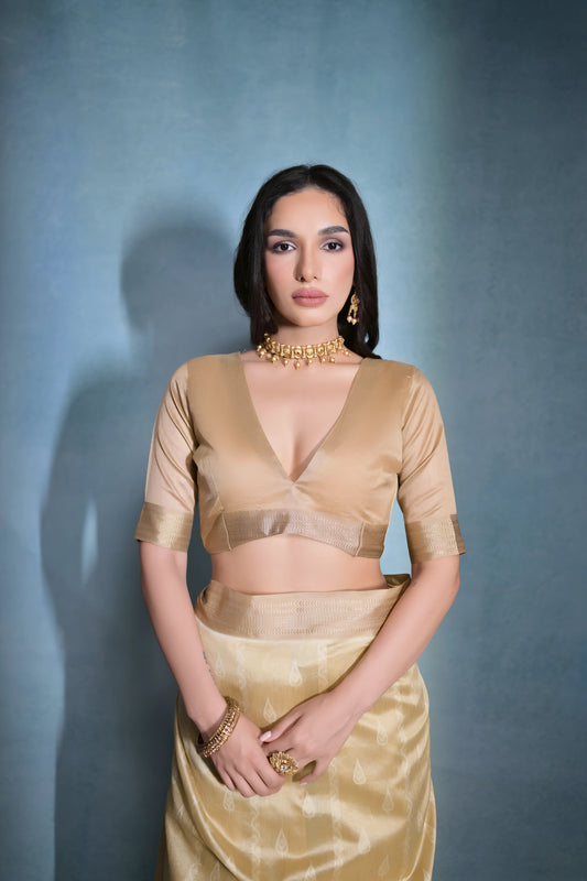 Woman wearing a golden silk saree with intricate border patterns and matching blouse, adorned with traditional gold jewelry including a choker necklace and earrings, posing elegantly in a studio setting with a soft blue gradient background.