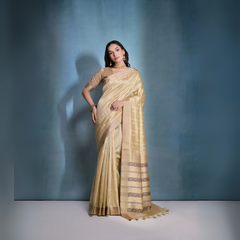 Woman wearing a golden silk saree with intricate border patterns and matching blouse, adorned with traditional gold jewelry including a choker necklace and earrings, posing elegantly in a studio setting with a soft blue gradient background.