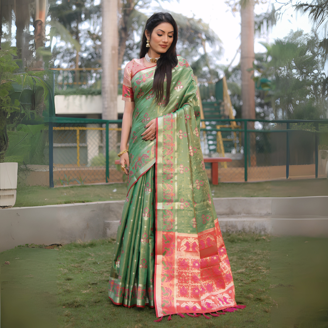 Woman wearing a green Banarasi silk saree with gold zari motifs and a vibrant contrast pallu featuring golden temple and pink floral patterns, paired with a short sleeve pink blouse and traditional gold jewelry. She stands elegantly outdoors on a grassy lawn surrounded by landscaped garden features, fencing, and tall tropical trees, creating a graceful natural setting.
