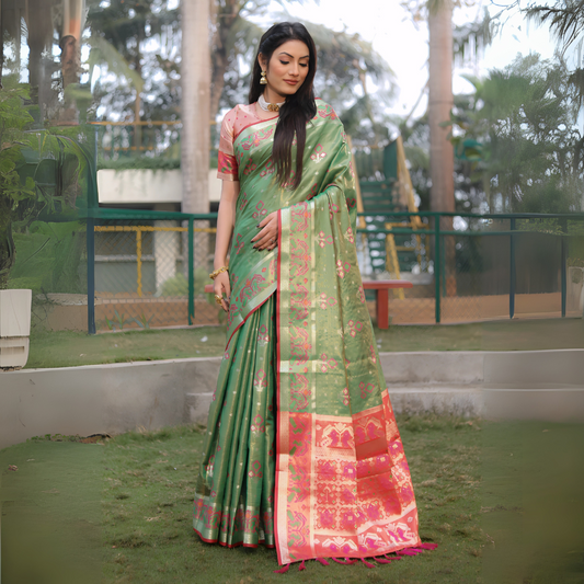Woman wearing a green Banarasi silk saree with gold zari motifs and a vibrant contrast pallu featuring golden temple and pink floral patterns, paired with a short sleeve pink blouse and traditional gold jewelry. She stands elegantly outdoors on a grassy lawn surrounded by landscaped garden features, fencing, and tall tropical trees, creating a graceful natural setting.