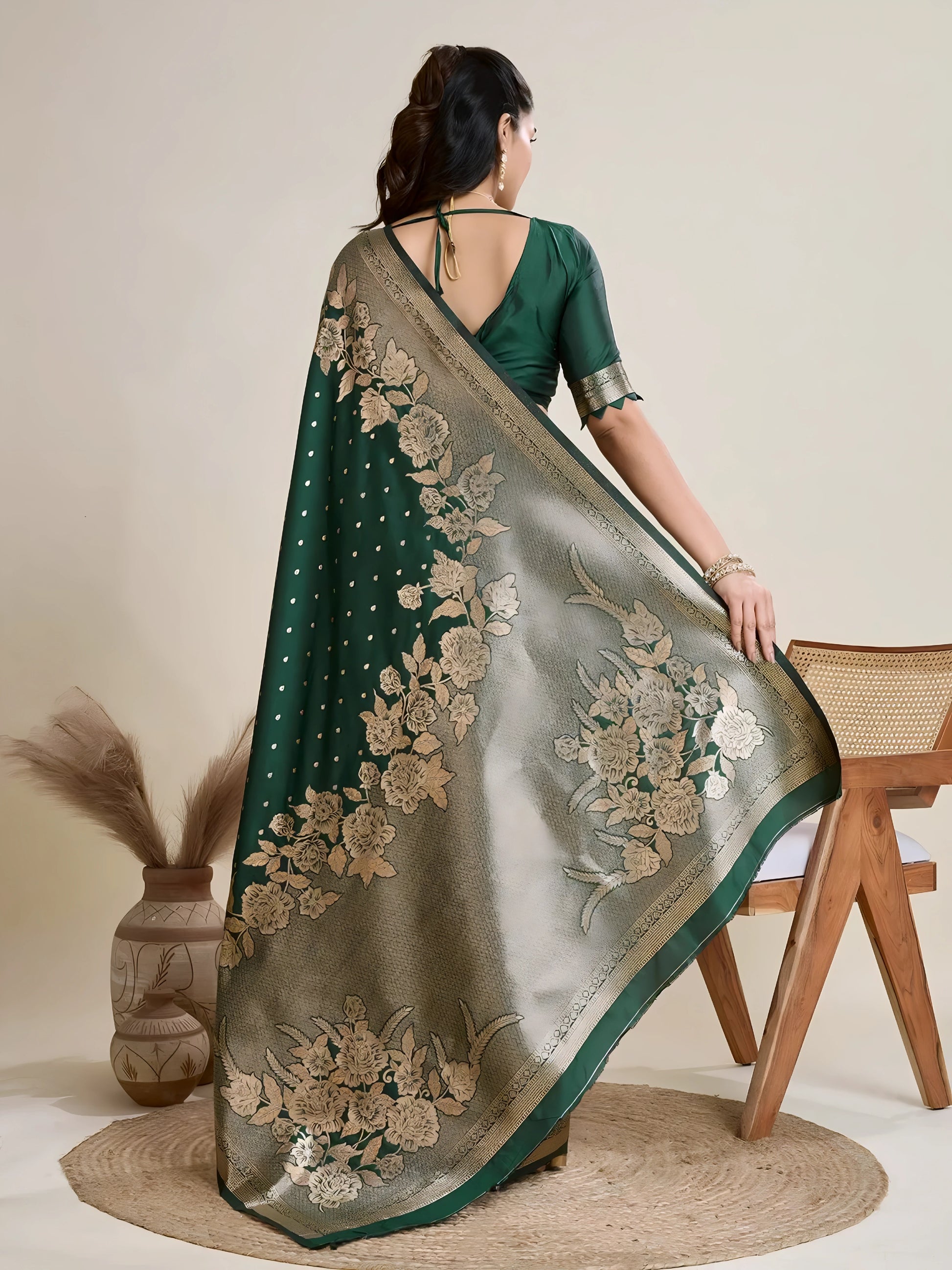 Woman wearing a green Banarasi silk saree with intricate gold floral zari border and pallu, paired with a matching blouse and traditional jewelry, elegantly posing beside a wooden chair in a minimalist studio with neutral decor, ceramic vases, and pampas grass.