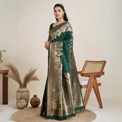 Woman wearing a green Banarasi silk saree with intricate gold floral zari border and pallu, paired with a matching blouse and traditional jewelry, elegantly posing beside a wooden chair in a minimalist studio with neutral decor, ceramic vases, and pampas grass.