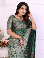 Woman wearing a green Banarasi silk saree adorned with intricate silver zari border and traditional woven motifs, paired with a matching silver blouse and classic jewelry. She poses gracefully in an elegant white room with decorative wall panels, white vases, and abundant pastel floral arrangements.