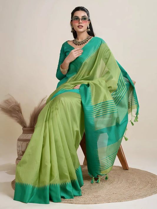 Woman wearing a green dual-tone silk saree with a contrasting teal blue border and pallu, paired with a matching green blouse and traditional jewelry, elegantly posing beside a wooden chair in a minimalist studio setting with neutral decor, ceramic vases, and pampas grass.