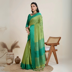Woman wearing a green dual-tone silk saree with a contrasting teal blue border and pallu, paired with a matching green blouse and traditional jewelry, elegantly posing beside a wooden chair in a minimalist studio setting with neutral decor, ceramic vases, and pampas grass.