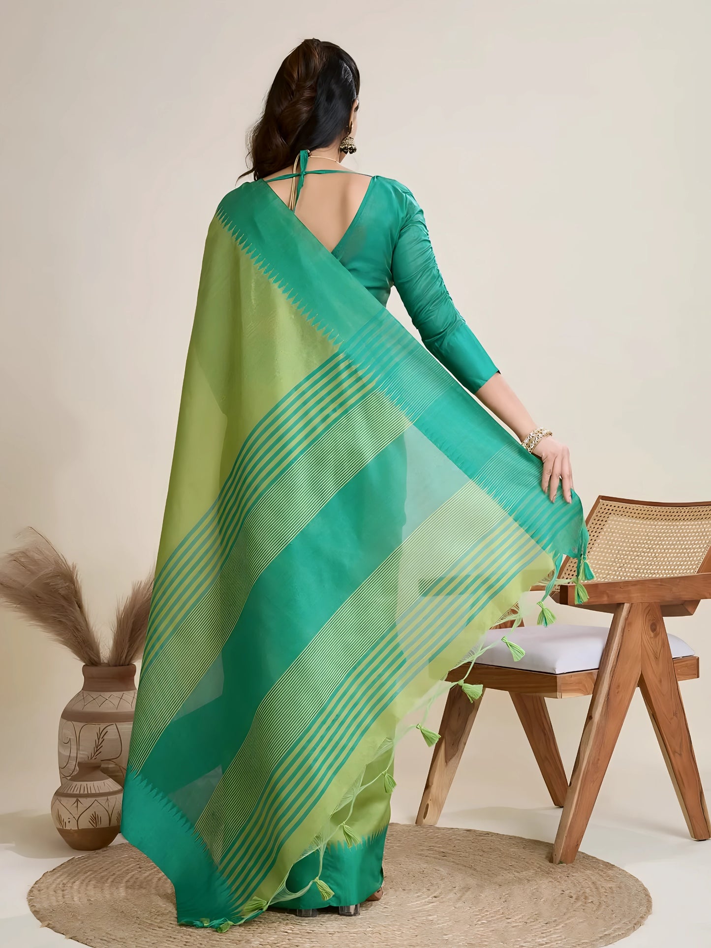Woman wearing a green dual-tone silk saree with a contrasting teal blue border and pallu, paired with a matching green blouse and traditional jewelry, elegantly posing beside a wooden chair in a minimalist studio setting with neutral decor, ceramic vases, and pampas grass.


