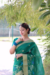 Woman wearing a green organza saree with floral embroidery and a light green border, posing outdoors under leafy trees on a bright pathway.