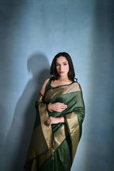 Woman wearing a green silk saree with gold border and traditional jewelry, posing against a plain blue background.