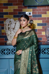 Woman wearing an elegant green silk saree with golden zari floral motifs and a rich gold border, accessorized with traditional gold jewelry, gracefully posing in a stylish vintage-inspired room with a blue cabinet, multicolored brick wall, decorative art, and a patterned armchair.