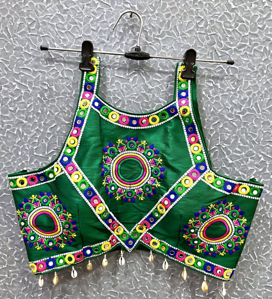 Green blouse featuring vibrant circular and oval mirror work with traditional Kutch multicolor embroidery, accented by geometric borders and shell tassel drops along the hem. Displayed on a hanger against a textured silver background, perfectly showcasing its festive and culturally inspired ethnic design.