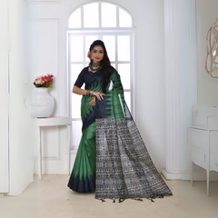 Woman wearing a green and navy blue Ikat printed saree with geometric black and white pallu, paired with a navy blouse, standing in a modern white interior with a large window and a vase of pink flowers on a console table.