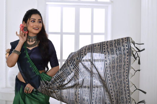 Woman wearing a green and navy blue Ikat printed saree with geometric black and white pallu, paired with a navy blouse, standing in a modern white interior with a large window and a vase of pink flowers on a console table.