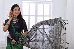 Woman wearing a green and navy blue Ikat printed saree with geometric black and white pallu, paired with a navy blouse, standing in a modern white interior with a large window and a vase of pink flowers on a console table.