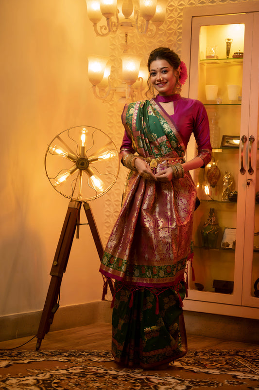 Woman wearing a green Banarasi silk saree with pink and gold floral zari border and pallu, paired with a magenta blouse and traditional bangles, elegantly seated on a white and pink armchair in a warmly lit vintage interior with a chandelier, decorative lamp, and glass display cabinet.