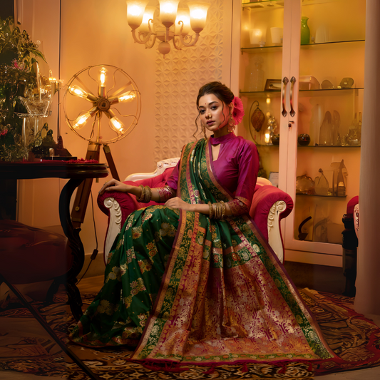 Woman wearing a green Banarasi silk saree with pink and gold floral zari border and pallu, paired with a magenta blouse and traditional bangles, elegantly seated on a white and pink armchair in a warmly lit vintage interior with a chandelier, decorative lamp, and glass display cabinet.