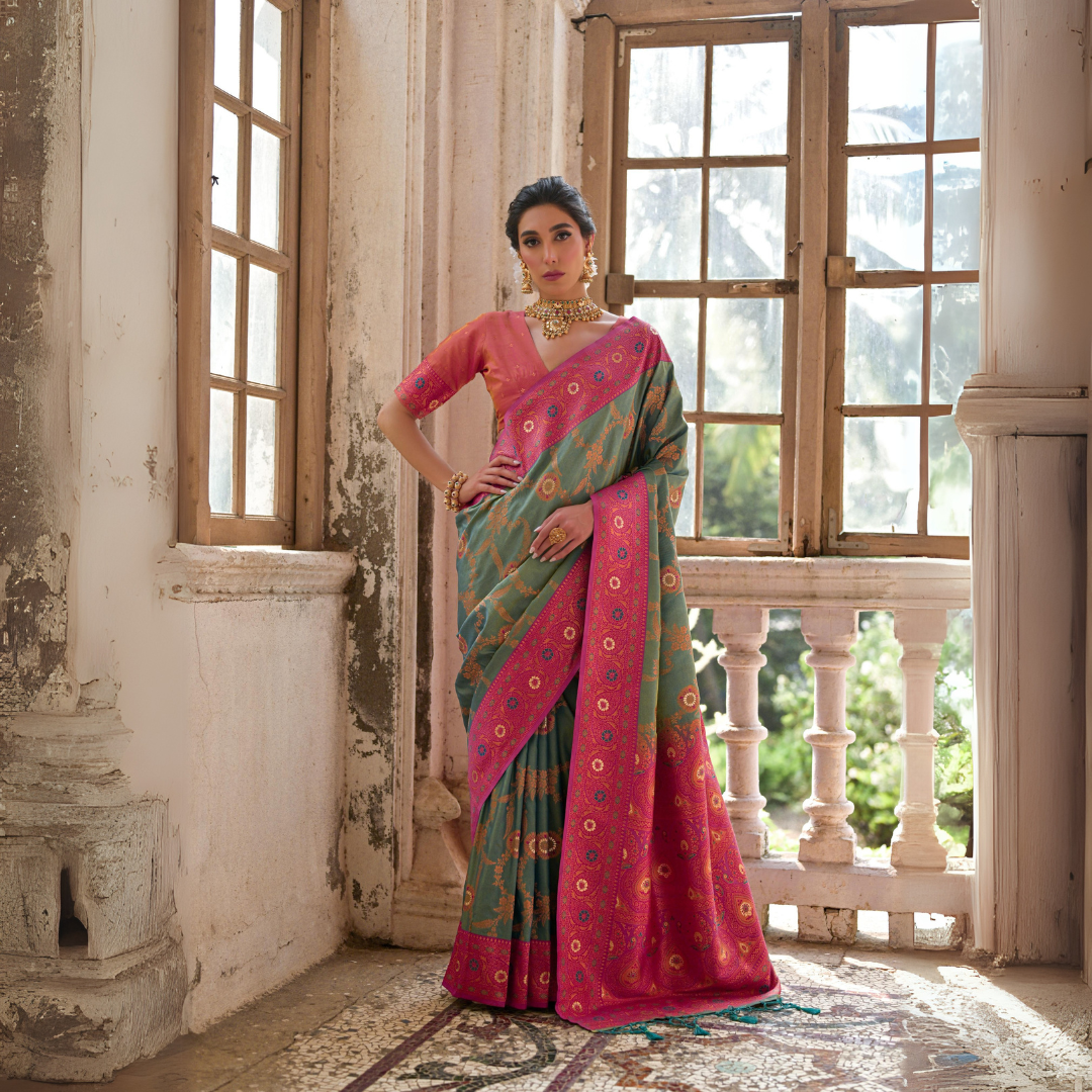 Woman wearing a green silk saree with intricate pink floral zari border and circular motifs, paired with a matching pink blouse and traditional gold jewelry, standing gracefully in a vintage room with large antique wooden windows and ornate architectural details.