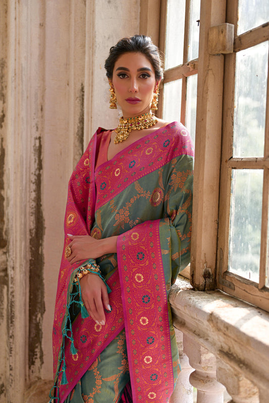 Woman wearing a green silk saree with intricate pink floral zari border and circular motifs, paired with a matching pink blouse and traditional gold jewelry, standing gracefully in a vintage room with large antique wooden windows and ornate architectural details.