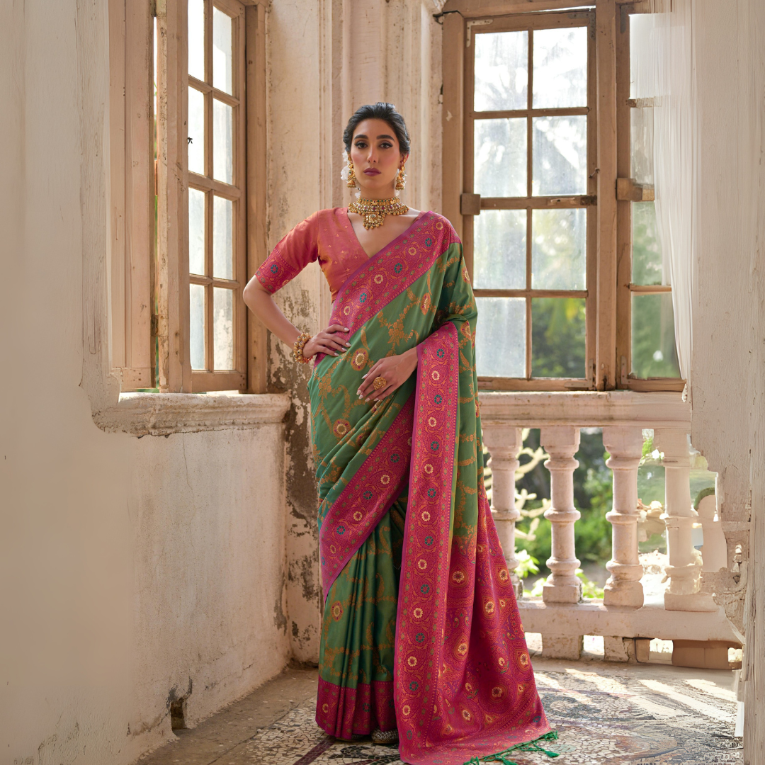 Woman wearing a green silk saree with intricate pink floral zari border and circular motifs, paired with a matching pink blouse and traditional gold jewelry, standing gracefully in a sunlit vintage room with large antique wooden windows and ornate architectural details.